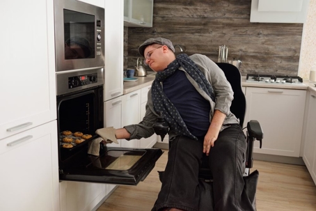 Man with disability using wheelchair taking homemade cookies out of electric oven in kitchen