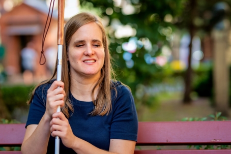 Visually impaired woman holds white cane and sits on bench in a city