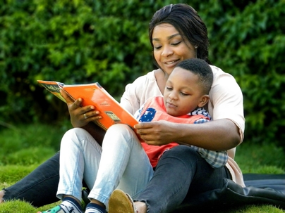 A woman and child are reading a book together outside. The image is likely a stock photo intended for use on a website.