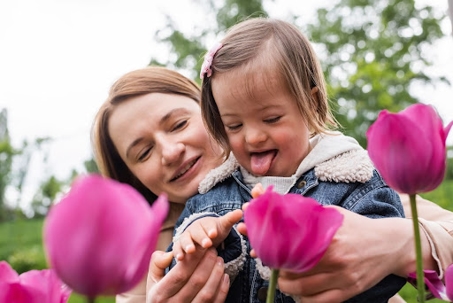 A woman embraces a child with Down syndrome, who playfully touches a pink tulip.